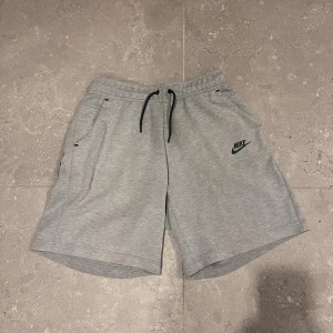 Grå Nike tech shorts  - Snygga grå shorts från Nike med svart logga på vänster ben. De har elastisk midja med svart snörning och praktiska fickor på sidorna. Perfekta för träning eller chill, tillverkade i mjukt och lätt material som känns skönt mot huden.