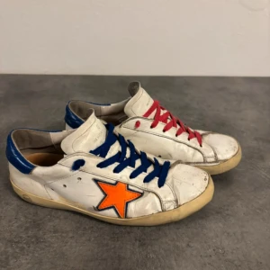 Golden Goose sneakers med färgglada detaljer - Unika Golden Goose sneakers i vitt skinn med blå och röda snören, blå häl och en orange stjärna på sidan. Platt sula och rund tå ger en klassisk look, medan de färgglada detaljerna gör dem riktigt iögonfallande. Perfekta för dig som gillar att sticka ut med din stil.