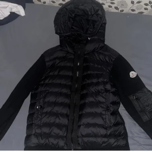 Svart Moncler cardigan  - Svart pufferjacka från Moncler med stickade ärmar och huva. Jackan har dragkedja framtill, två fickor med dragkedja och Moncler-logga på vänster ärm. Kroppen är quiltad och tillverkad i syntetmaterial medan ärmarna är stickade för extra komfort.