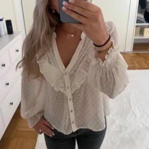 Vit blus med volang och prickar - Söt vit blus med volangdetaljer och små prickar. Blusen har v-ringning, knappar framtill och långa puffiga ärmar med resår vid handlederna. Tunt, luftigt material som ger en romantisk vibe.