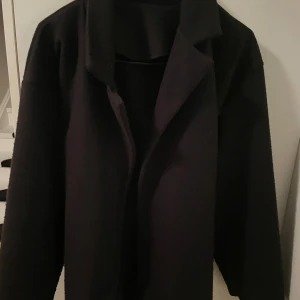 Svart kappa ifrån missay  - Svart kappa med klassisk krage och bälte i midjan. Jackan har lång ärm och en stilren, rak passform. Materialet ser ut att vara ull eller liknande, perfekt för kyliga dagar. Enkel och snygg design som funkar till många olika outfits.