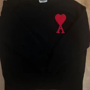 Svart stickad tröja från Ami med hjärta - Svart stickad tröja från Ami med ett stort rött hjärta och bokstaven A broderat på bröstet. Rund hals och långa ärmar med ribbade muddar. Perfekt för dig som gillar stilrena plagg med en cool detalj.