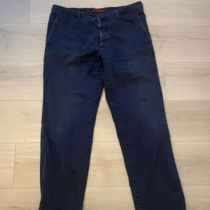 Prada Mörkblå chinos  - Snygga mörkblå chinos från Prada med klassisk rak passform och diskret röd detalj på insidan av midjan. Byxorna har dragkedja med metallzip där det står Prada Milano. Tillverkade i mjuk bomull med fickor fram och bak.