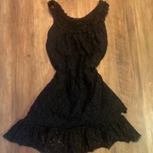 Svart spetsklänning med volang - Supersnygg svart kortklänning med spets och volangkant längst ner. Klänningen är ärmlös och har en feminin, figurnära passform. Perfekt för dig som gillar romantiska detaljer och vill sticka ut med en cool look.