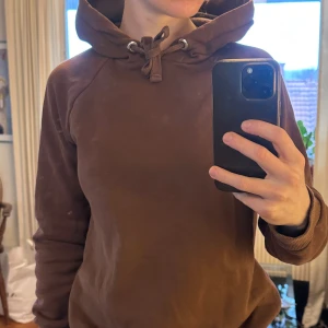 Brun hoodie från Gina Tricot - Mysig brun hoodie från Gina Tricot i storlek S. Tröjan har huva med snörning och är tillverkad i mjuk fleece på insidan. Klassisk passform med långa ärmar och ribbade muddar vid ärmslut och nederkant. 