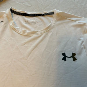 Under Armour vit/grå tränings-tshirt - Snygg och sportig t-shirt från Under Armour i vit och ljusgrå färg. T-shirten har rund hals, korta ärmar och är tillverkad i lätt polyester som andas. Perfekt för löpning eller gym, med UA-logga på bröstet och coola detaljer vid halsen.