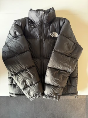 Svart North Face 1996 Retro  - Svart pufferjacka från The North Face i storlek M. Jackan har en hög krage, två sidofickor med dragkedja och en innerficka med zip. Materialet är syntet och dun, perfekt för kalla dagar. Klassisk logga på bröstet och insidan, även på baksidan! 
