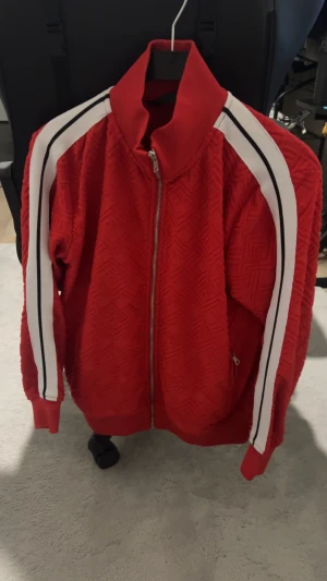 Röd kofta från Palm Angels - Palm Angels tröja i rött med vit och svart rand längs ärmarna. tröjan har ett snyggt geometriskt mönster i tyget, dragkedja framtill och ståkrage. Materialet känns som polyester och passformen är normal. Perfekt för dig som gillar streetwear.