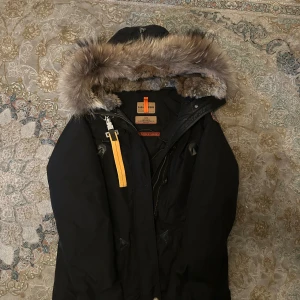 Svart Parajumpers parkas med päls - Svart parkas från Parajumpers med stor huva och fluffig päls i beige/bruna toner. Jackan har quiltad insida, dragkedja, knappar och en gul detaljrem. Märkespatch på ryggen och flera praktiska fickor. Perfekt för kalla vinterdagar. Bara använd två gånger kvitto finns