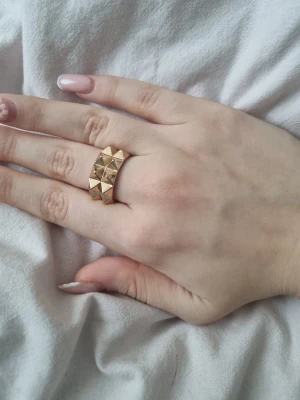 Guldfärgad ring med pyramidmönster - Säljer en cool guldfärgad ring med upphöjda pyramidformade detaljer runt hela ringen. Den har en chunky och edgy vibe som verkligen sticker ut. Perfekt för dig som gillar statement-smycken och vill addera lite attitude till din stil.