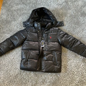 Svart pufferjacka från Polo Ralph Lauren - Svart pufferjacka från Polo Ralph Lauren med huva, dragkedja och knappstängning. Jackan har en röd broderad logga på bröstet, flera fickor och en glansig finish. Perfekt för kalla dagar och har en riktigt clean look. Pris kan diskuteras 