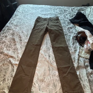 Olivgröna chinos från Dressmann - Säljer ett par olivgröna chinos från Dressmann med klassisk rak passform. Byxorna har normal midja och är tillverkade i mjuk bomull, perfekta för en avslappnad men ändå stilren look.