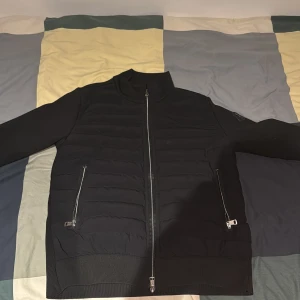 Svart pufferjacka från Moncler Grenoble - Svart pufferjacka från Moncler Grenoble med quiltad front, två dragkedjeförsedda fickor och ribbade detaljer på ärmar och nederkant. Jackan har hög krage och Moncler-logga på vänster ärm. Materialet är polyester och syntet, perfekt för kyliga dagar.