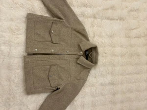 Beige jacka från ZARA - Säljer en beige jack från ZARA i storlek S. Jackan har klassisk krage, stora fickor med lock och silverfärgade knappar framtill. Materialet är mjukt och känns som ull, perfekt för lager-på-lager. Enkel och clean design som funkar till många stilar.
