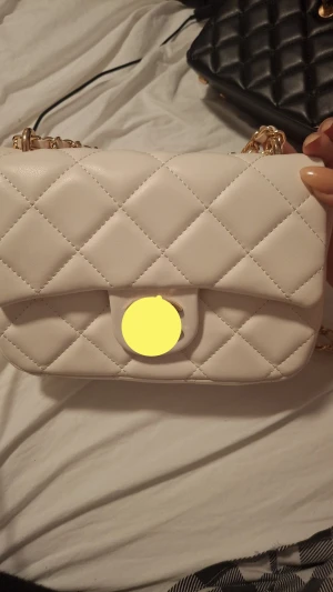 Vit quiltad axelväska med guldkedja - Supersnygg vit axelväska med quiltat mönster och guldfärgad kedja som axelrem. Väskan har en stilren design med rundade kanter och lock med knäppning framtill. Perfekt för dig som vill ha en trendig och elegant accessoar.