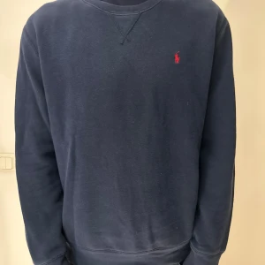 Marinblå sweatshirt från Ralph Lauren - Snygg marinblå sweatshirt från Ralph Lauren med klassisk röd logga broderad på bröstet. Tröjan har rund hals, långa ärmar och ribbade muddar vid ärmslut och nederkant. Perfekt för dig som gillar stilrena och tidlösa plagg.