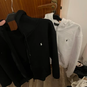 Svart och vit hoodie från Polo Ralph Lauren - Två hoodies från Polo Ralph Lauren, en svart och en vit, båda med klassisk broderad logga på bröstet. Hoodiesarna har huva, snörning och dragkedja framtill. Perfekt för dig som gillar stilren och enkel design. Svart använd 1 gång, annars nya. A kopia 