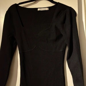 Svart ribbad långklänning från Adoore - Svart långklänning från Adoore med ribbad struktur och figurnära passform. Klänningen har långa ärmar, markerad byst och en snygg, något djupare urringning. Perfekt för dig som gillar stilrena och tidlösa plagg med en modern twist.