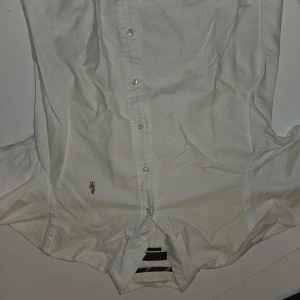 Vit skjorta från Polo Ralph Lauren - Vit klassisk skjorta från Polo Ralph Lauren med broderad logga på bröstet. Skjortan har knappar framtill och lång ärm, perfekt för en clean och stilren look. Materialet är mjuk bomull som känns skönt mot huden.