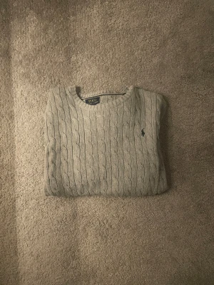 Grå stickad tröja från Polo Ralph Lauren - Snygg grå stickad tröja från Polo Ralph Lauren med klassiskt kabelstickat mönster och rund hals. Tröjan har en liten broderad logga på bröstet och är perfekt för dig som gillar stilrena och tidlösa plagg. Passar dig som vill ha en chill och clean look.