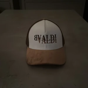 Brun och vit keps från BVALDI - Snygg truckerkeps från BVALDI med vit frontpanel och brunt nät baktill. Skärmen är beige och kepsen har ett coolt BVALDI-tryck framtill. Perfekt för dig som vill ha en stilren och trendig accessoar. Justerbar passform och ventilerande meshmaterial.