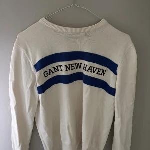 Vit stickad tröja GANT New Haven - Vit stickad tröja från GANT med blåa ränder och texten 'GANT NEW HAVEN' över bröstet. Tröjan har rund halsringning och långa ärmar. Perfekt för dig som gillar klassisk och sportig stil med en touch av college-vibe.