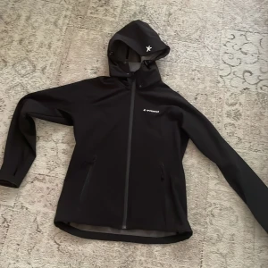 Svart vindjacka från Everest med huva - Svart vindjacka från Everest med stilren design och huva. Jackan har dragkedja framtill, två sidofickor med dragkedja och diskret logga på bröstet och huvan. Perfekt för blåsiga dagar och outdoor-aktiviteter.