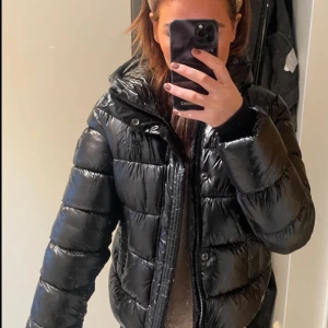 Svart pufferjacka från Superdry - Säljer en svart, glansig pufferjacka med huva och dragkedja. Jackan är vadderad och har en oversized look, perfekt för kalla dagar. Den har långa ärmar och praktiska fickor framtill. Passar dig som vill ha en stilren och trendig vinterjacka.