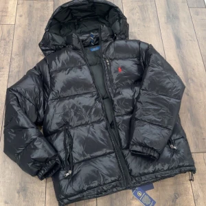  Polo Ralph Lauren jacka - Svart pufferjacka från Polo Ralph Lauren med huva och dragkedja. Jackan har en glansig finish, två sidofickor med dragkedja och röd broderad logga på bröstet. Justerbar nederkant med snörning och quiltad design för extra värme. Perfekt för kalla dagar.