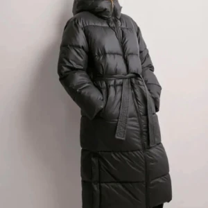 Svart lång pufferjacka med bälte - Säljer en svart, lång pufferjacka med huva och bälte i midjan. Jackan har quiltad design och dragkedja framtill. Perfekt för kalla dagar när du vill hålla dig varm och samtidigt se cool ut. Jackan har fickor på sidorna och är medel lång.