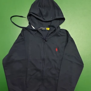 Mörkblå hoodie från Polo Ralph Lauren - Snygg mörkblå hoodie från Polo Ralph Lauren med röd broderad logga på bröstet. Tröjan har huva med snörning, dragkedja framtill och långa ärmar. Tillverkad i mjuk bomull för skön komfort. Perfekt för chill dagar och streetstyle.