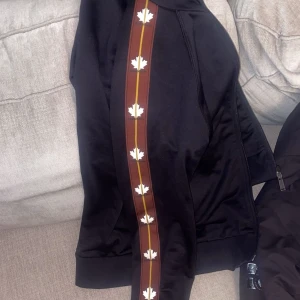 Dsquared2 kofta - Svart hoodie från Dsquared2 med bruna och gula ränder längs ärmarna, dekorerade med vita lönnlöv. Tröjan har huva, dragkedja och ribbade muddar. Materialet är mjuk polyester som ger en skön känsla. Perfekt för dig som gillar streetwear med coola detaljer.