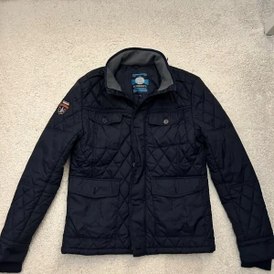 Mörkblå quiltad jacka från Bendoff - Snygg mörkblå quiltad jacka från Bendoff med krage och flera fickor framtill. Jackan har ett marin-inspirerat märke på ärmen och knappar på fickorna. Perfekt för dig som gillar stilren och sportig look. S passar M