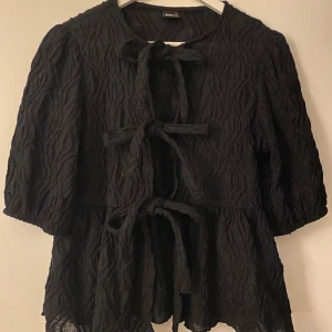 Svart blus med knyt från Gina Tricot - Svart blus från Gina Tricot med strukturerat tyg och tre knytningar framtill. Modellen har halvlånga puffärmar och lätt volang nertill. Perfekt för dig som gillar unika detaljer och en trendig look.