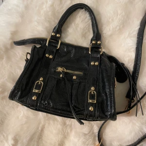 Svart handväska med gulddetaljer - Snygg svart handväska i glansigt skinn och guld detaljer. Köpte på någon sellout butik med väskor i Nice, står inget märke så ingen aning om kvalitet då jag har inte fått någon användning av den. Köpte den för 330 kr