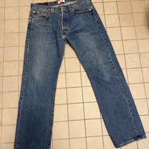 Levi's 501 blå jeans straight fit - Klassiska Levi's 501 jeans i blå denim med raka ben och fem fickor. Jeansen har knappgylf, kontrastsömmar och den ikoniska läderpatchen bak i midjan. Perfekt för dig som gillar tidlös stil och vill ha ett par jeans som alltid funkar.