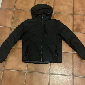 Svart pufferjacka från Polo Ralph Lauren - Svart pufferjacka från Polo Ralph Lauren med huva och röd broderad logga på bröstet. Jackan har dragkedja framtill och är tillverkad i syntetmaterial. Perfekt för kalla dagar och har en clean, modern look.
