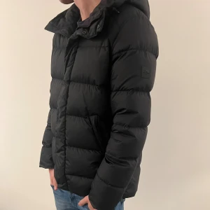 Svart pufferjacka från J.Lindeberg - Svart pufferjacka från J.Lindeberg med huva och diskret logotyp på ärmen. Jackan har en quiltad design, dragkedja och knappar framtill samt två sidofickor. Perfekt för kalla dagar och har en clean, modern look som funkar till jeans och sneakers.