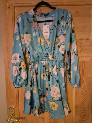 Blommig klänning från Zara - Säljer en ljusblå kort omlottklänning från Zara med v-ringning och blommigt mönster i rosa, gul och svart. Klänningen har långa ärmar med resår vid handlederna, volang nedtill och knyt i midjan för en snygg siluett. Perfekt för dig som gillar färg och mönster! Aldrig använd, prislapp kvar.