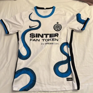 Inter Milan vit matchtröja Nike - Säljer en Inter Milan fotbollströja från Nike i vit polyester med blå och svart ormmönster. Kortärmad modell med svart och blå detaljer på krage och ärmslut. Tryck med $INTER Fan Token och Lenovo på ryggen. Perfekt för dig som älskar Serie A och snygga matchtröjor.