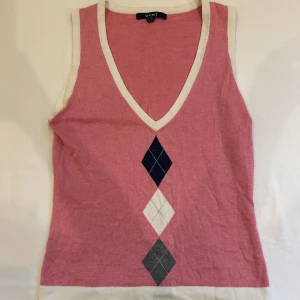 Rosa argyle väst från GANT - Snygg rosa vintage argyle väst från GANT med v-ringning och vita kanter. Framsidan har klassiskt argylemönster i mörkblått, vitt och grått. Toppen är ärmlös och gjord i mjuk bomull. 