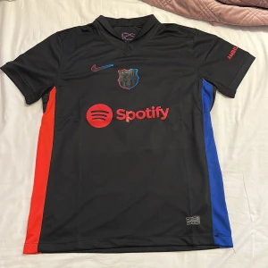FC Barcelona svart matchtröja Nike - Säljer en FC Barcelona fotbollströja med Lamine Yamal och nummer 10 på ryggen. Tröjan är svart med röda och blå sidopaneler, röda tryck och detaljer, samt Spotify-logga på bröstet. Märkt med Nike och klubbens emblem. Materialet är lätt och andas, perfekt för träning eller match.