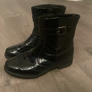 Svarta boots från Aaltonen med spänne - Snygga svarta boots från Aaltonen med krokodilpräglad yta och dekorativt spänne på sidan. Skorna har platt sula med extra grepp och dragkedja på insidan för enkel påtagning. Perfekt för dig som gillar stilrena och coola detaljer. Använda 1 gång. 