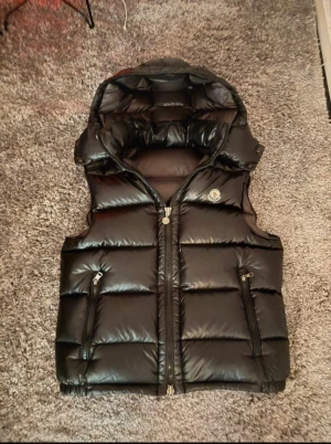 Svart dunväst från Moncler med huva storlek M, Pris kan diskuteras! - Snygg svart dunväst från Moncler med glansig finish och stor, vadderad huva. Västen har två dragkedjefickor fram, quiltad design och Moncler-logga på bröstet. Perfekt för lager-på-lager och streetstyle. Dragkedja framtill för enkel stängning.