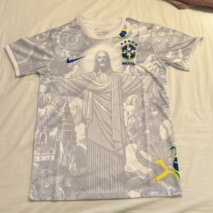 Vit Brasil Nike fotbollströja - Säljer en unik Brasil fotbollströja från Nike i vitt med detaljerat grafiskt tryck av Kristusstatyn och brasilianska byggnader. Kortärmad modell med blå Nike-logga och Brasil-märke på bröstet. Materialet är lätt och andas, perfekt för match eller träning.