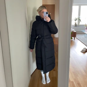Ny pufferjacka från Ellos, aldrig använd prislapp kvar  - Säljer en helt ny, lång pufferjacka från Ellos med huva och bälte i midjan. Jackan har quiltad design, dragkedja och knappar framtill samt långa ärmar. Perfekt för kalla dagar och riktigt snygg siluett med bältet i midjan. Bara testad med prislappen kvar. 