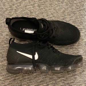 Nike Vapormax (str 42,5) - Säljer ett par svarta Nike Vapormax sneakers med vit swoosh. Skorna har stickad ovandel, snörning och genomskinlig bubblig sula som ger en futuristisk vibe. Perfekta för dig som gillar streetstyle och vill ha något bekvämt och snyggt.