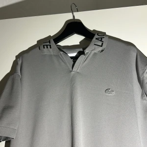 Grå pikétröja från Lacoste - Snygg grå pikétröja från Lacoste med klassisk krage där Lacoste-loggan är broderad i svart. Tröjan har korta ärmar och det ikoniska krokodilmärket på bröstet. Materialet är mjukt och känns lyxigt, perfekt för dig som gillar stilrena plagg.