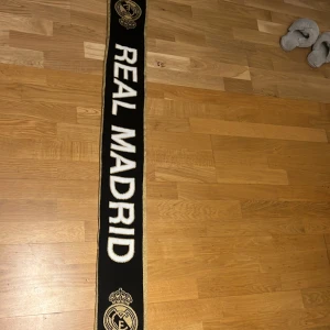 Real Madrid svart och guld halsduk - Snygg halsduk från Real Madrid i svart med guldiga detaljer och klubbens logga i båda ändar. Tjockt stickad med fransar i ändarna och stora vita bokstäver. Perfekt för att visa ditt stöd på matchen eller hänga upp på väggen.