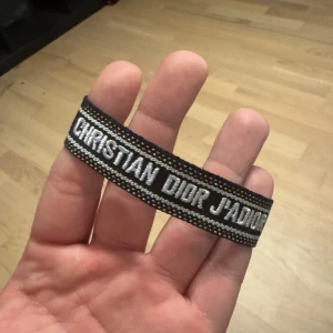 Christian Dior J'Adior svart tygbälte - Snyggt svart tygbälte från Christian Dior med texten 'J'Adior' broderad i vitt. Bältet har detaljer i guld och silver längs kanterna och är gjort i ett kraftigt vävt material. Perfekt accessoar för att lyfta din outfit med en lyxig touch.